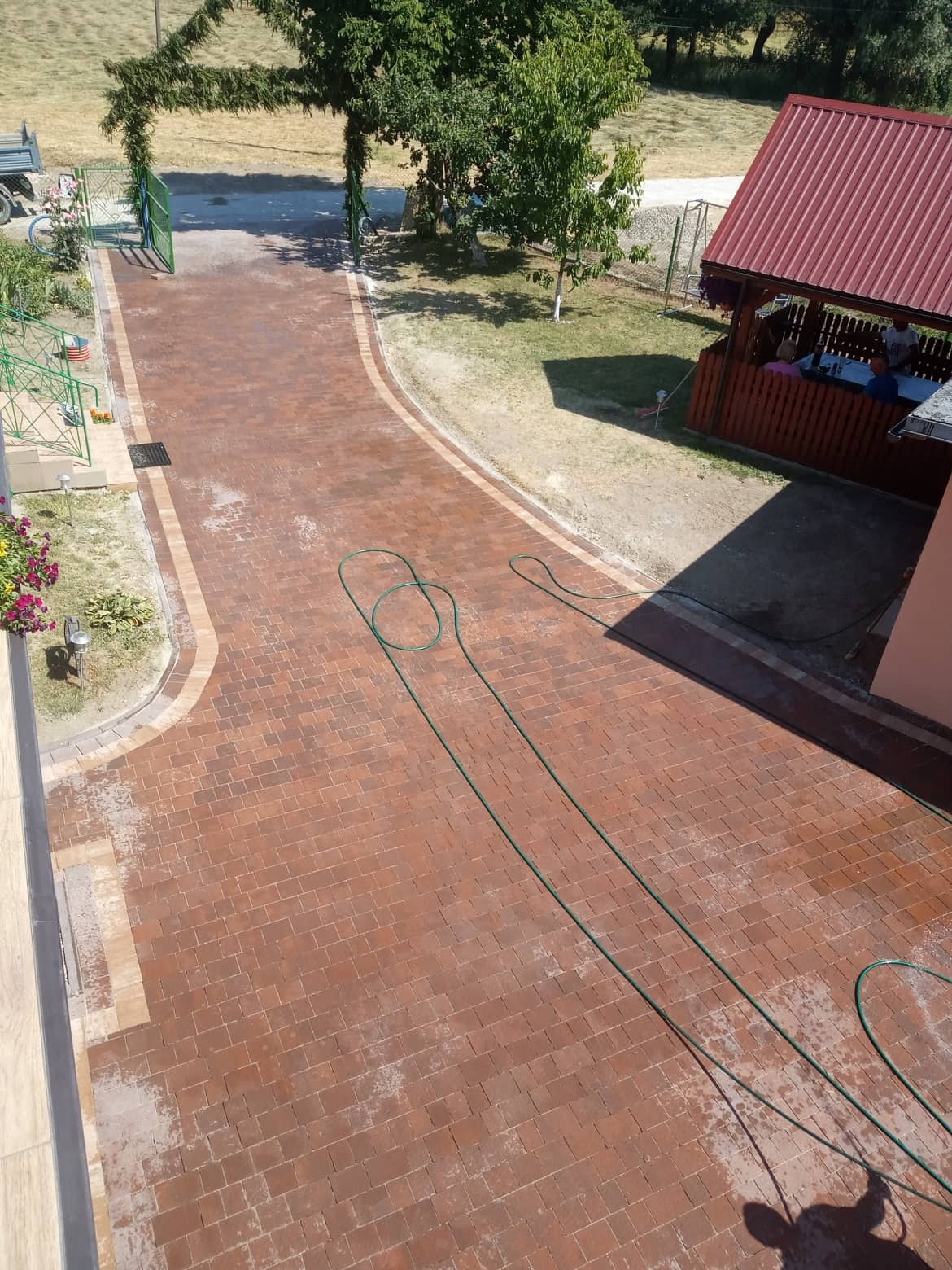 Widok z góry na nowo ułożoną, czerwoną kostkę brukową na podjeździe, z zielonym wężem ogrodowym leżącym na powierzchni. Z tyłu altana z ludźmi i brama z zieleni. Widoczne ślady mycia kostki.
