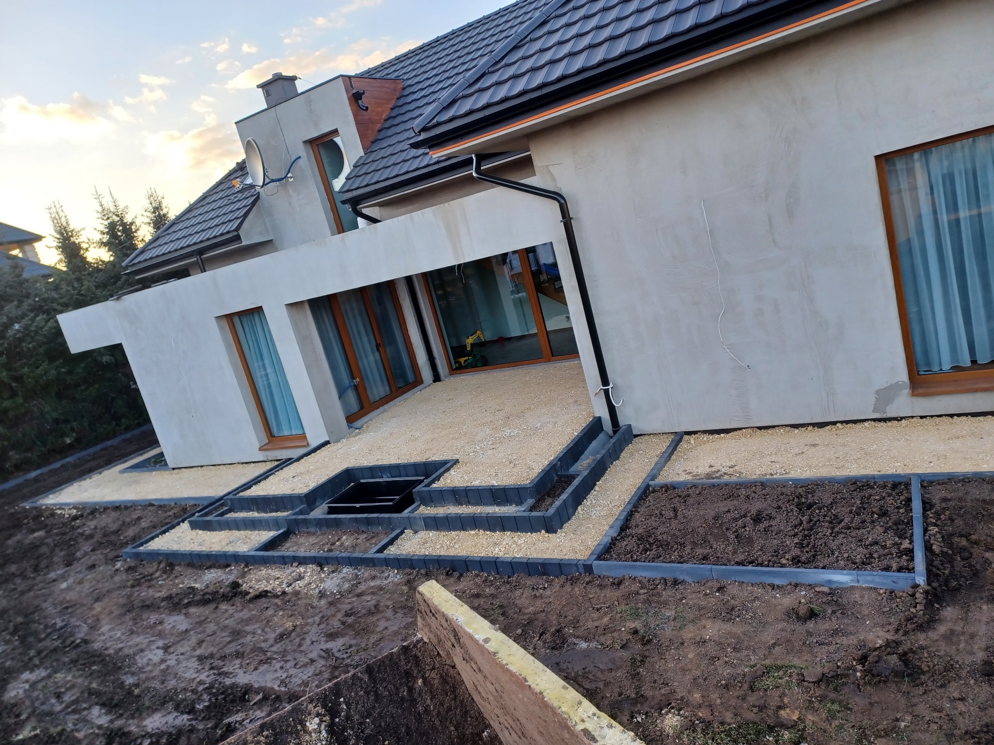 Taras domu jednorodzinnego z wysypanym żwirem, obrzeża z betonowych kostek, przygotowane rabaty ziemne, widok na elewację budynku z oknami.