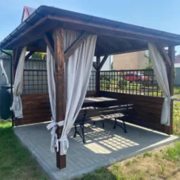 Drewniana konstrukcja z zadaszeniem i zasłonami w ogrodzie, wyposażona w stół i ławki, ustawiona na kostce brukowej, widoczny zbiornik na deszczówkę z tyłu.