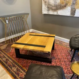 Kwadratowy stolik kawowy z jasnego drewna z czarnymi wstawkami, ustawiony na wzorzystym dywanie w salonie z designerską konsolą i abstrakcyjnym obrazem w tle.