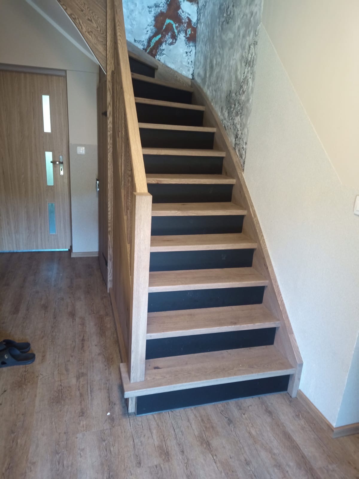 Nowoczesne schody z drewnianymi stopniami i czarnymi podstopnicami, widoczne z dołu, z drewnianą podłogą i drzwiami w tle, oraz dekoracyjną fakturą na ścianie.