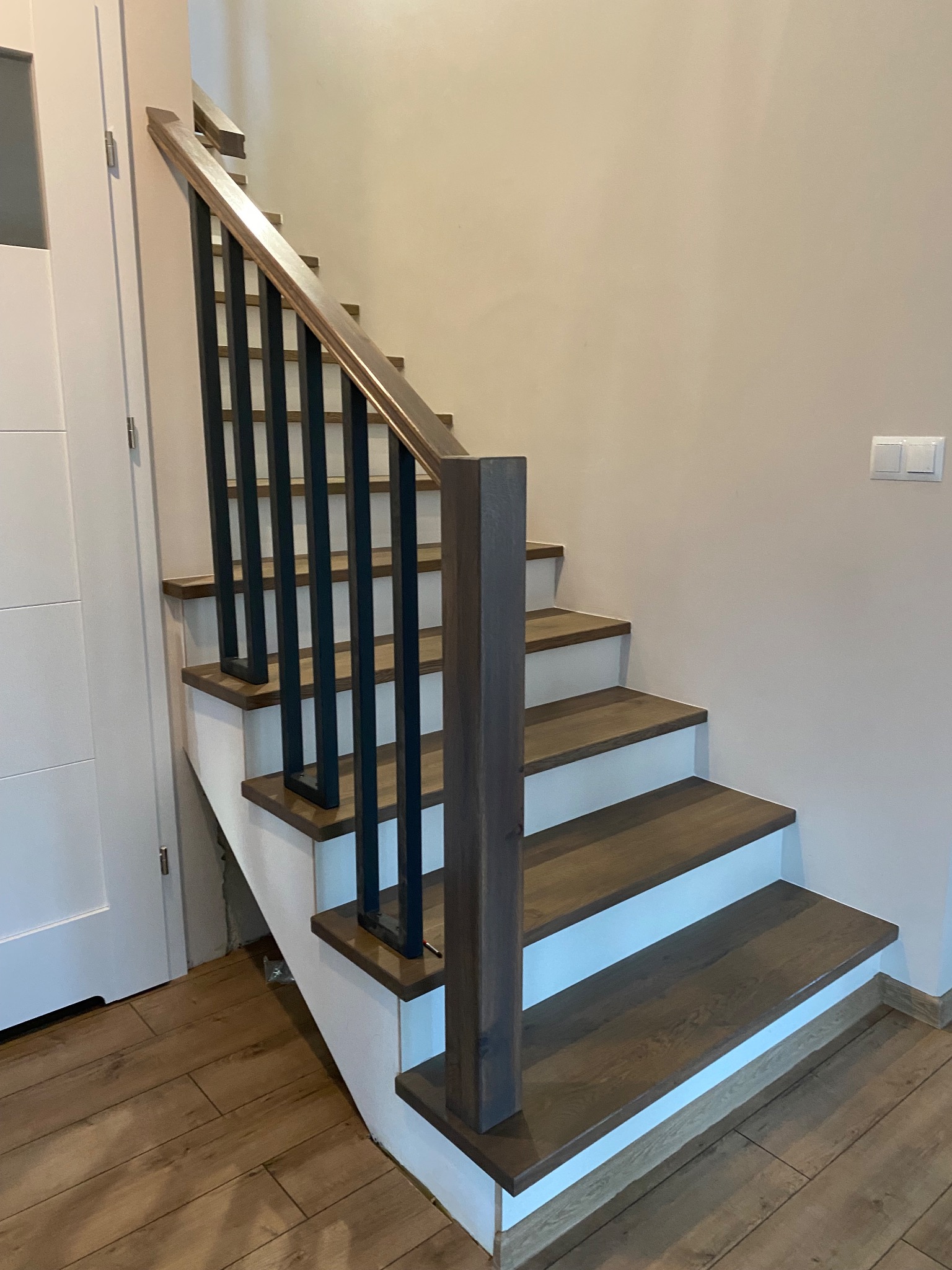 Nowoczesne schody drewniane z ciemnymi stopniami i czarnymi, prostymi balustradami, białe podstopnice, widok z dołu na górę.