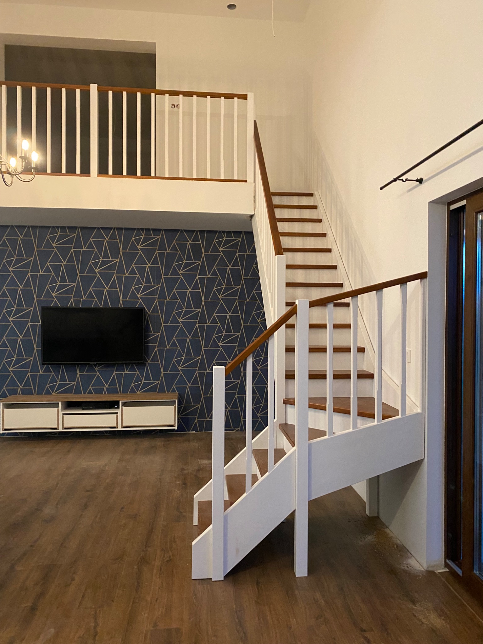 Dwubiegowe schody z drewnianymi stopniami i białymi balustradami w nowoczesnym wnętrzu z parkietem, na tle ściany z geometrycznym wzorem i telewizorem.