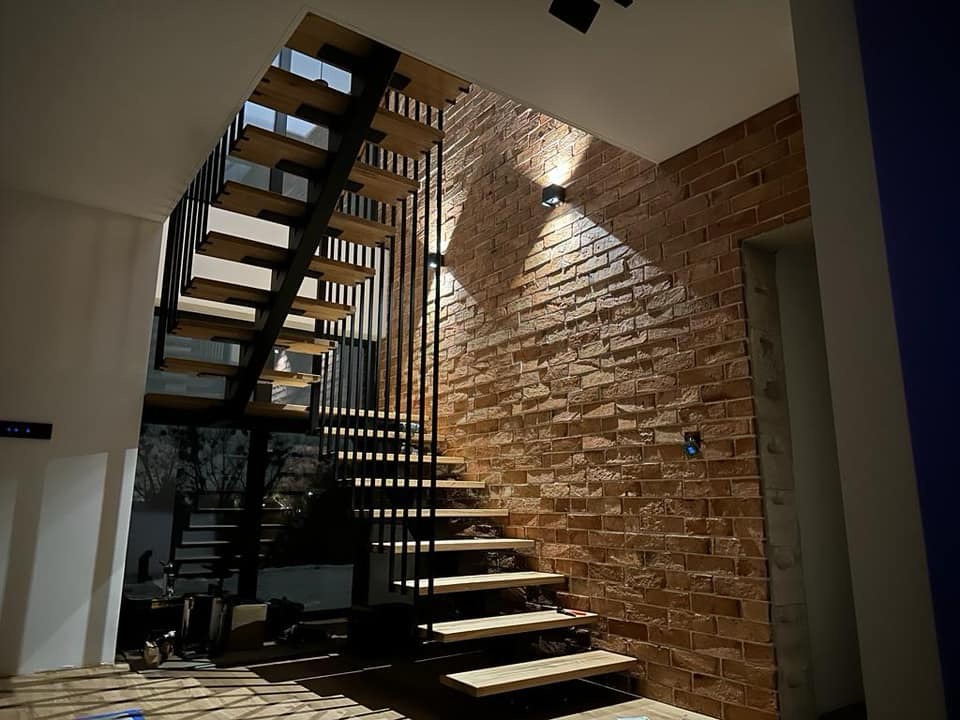Nowoczesne schody z drewnianymi stopniami i czarną metalową balustradą, ustawione przy ścianie z cegły, oświetlone punktowymi lampami.