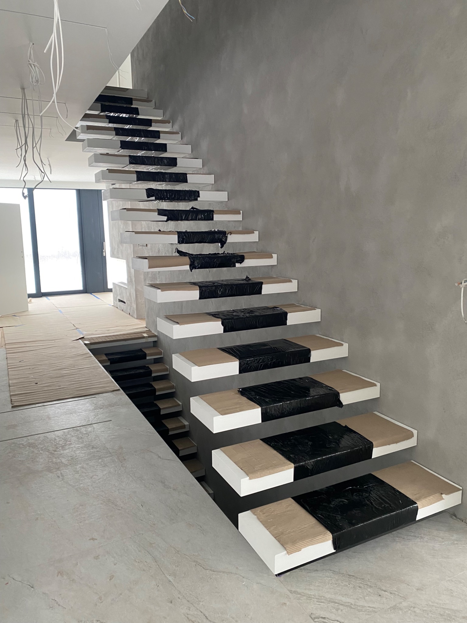 Nowoczesne schody wspornikowe z drewnianymi stopniami zabezpieczonymi folią, w surowym wnętrzu z szarą ścianą i widocznymi przewodami elektrycznymi.