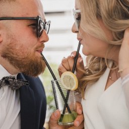 NATS PHOTOGRAPHY Natalia Głowacka - Para młoda w okularach pije drinka z dwóch słomek; panna młoda ma wpięte kwiaty we włosy, a pan młody ma muszkę.