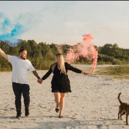 NATS PHOTOGRAPHY Natalia Głowacka - Para trzymająca się za ręce, biegnąca po piaszczystej plaży z kolorowymi dymami, z psem biegnącym obok, w tle drzewa.
