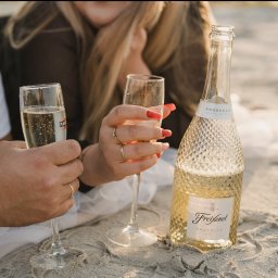 NATS PHOTOGRAPHY Natalia Głowacka - Para w plenerze na plaży, trzymająca kieliszki z Martini Prosecco i butelkę Freixenet Prosecco, z widocznymi pierścionkami na palcach i czerwonymi paznokciami u kobiety.