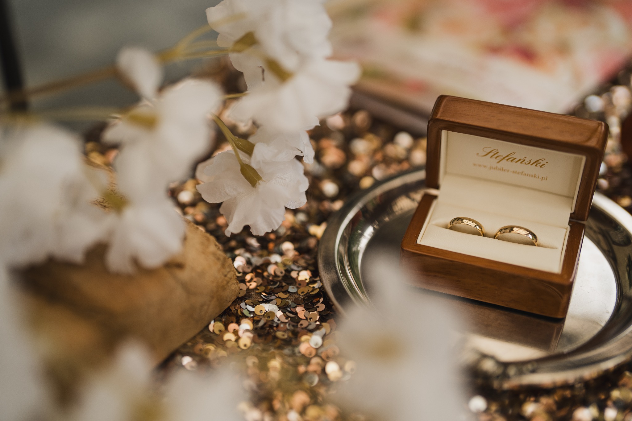 Dwa złote obrączki ślubne w drewnianym pudełku z logo jubilera Stefański, ustawione na srebrnej tacy z cekinami i dekorowane białymi kwiatami.