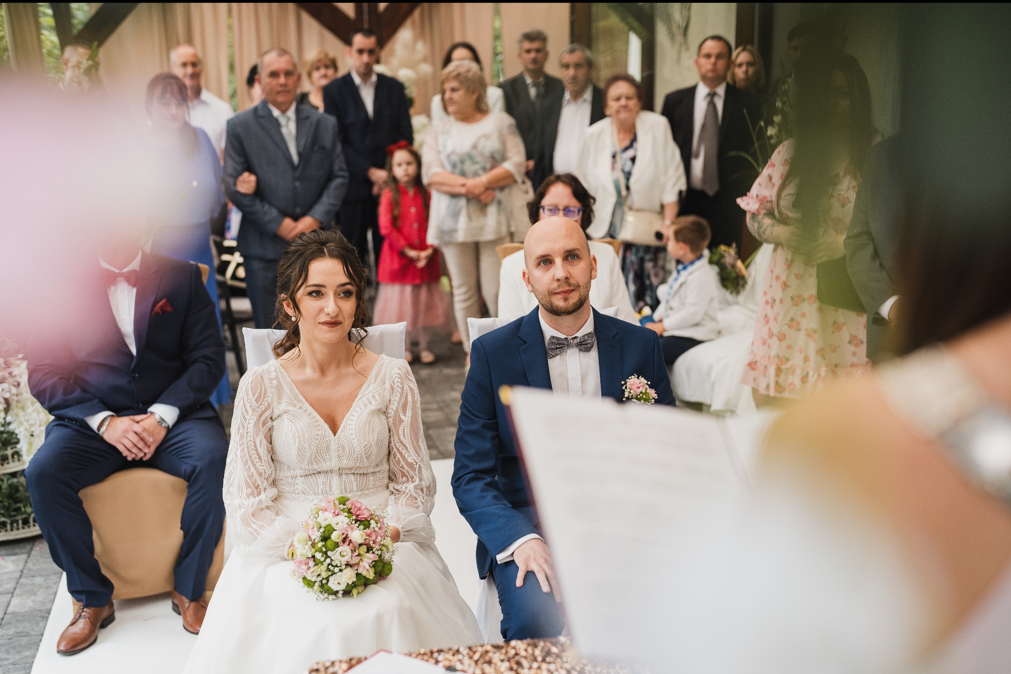 Para młoda siedzi podczas ceremonii ślubnej, panna młoda trzyma bukiet, a w tle widać gości.