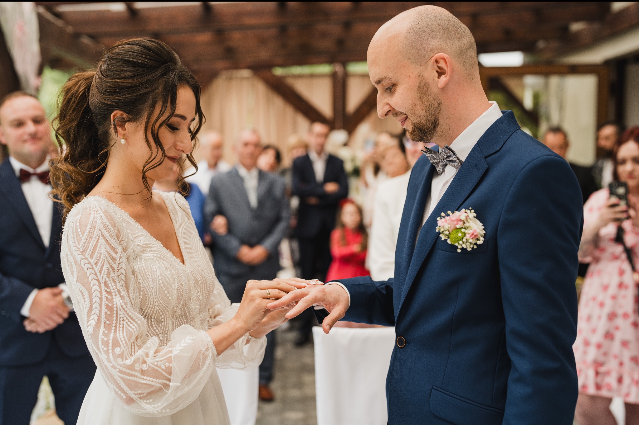 Panna młoda zakłada obrączkę na palec pana młodego podczas ceremonii ślubnej; w tle goście weselni i drewniana konstrukcja.