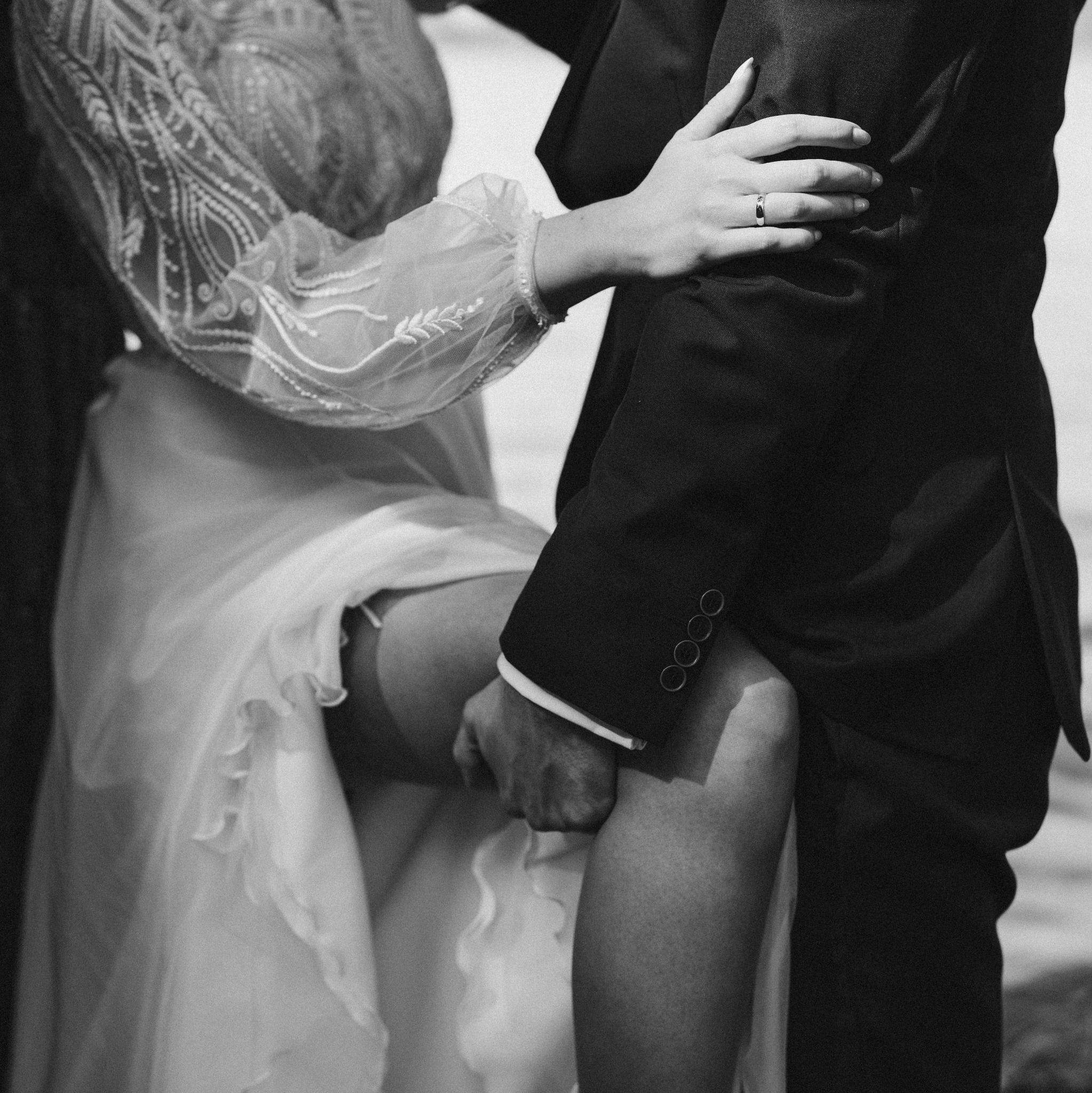 Czarno-białe zbliżenie pary młodej: panna młoda w koronkowej sukni i podwiązce, pan młody w czarnym garniturze, dłoń z obrączką na ramieniu.