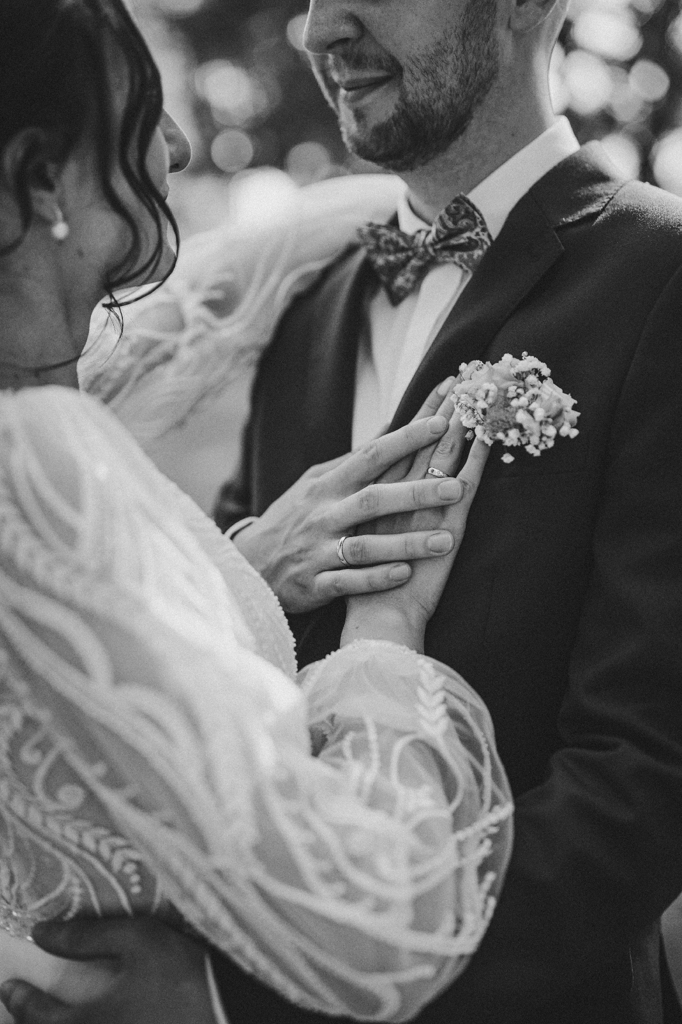 Czarno-białe zbliżenie dłoni pary młodej z obrączkami, bukiecik w butonierce pana młodego, wzorzysta mucha, fragment sukni ślubnej z koronkowymi detalami.