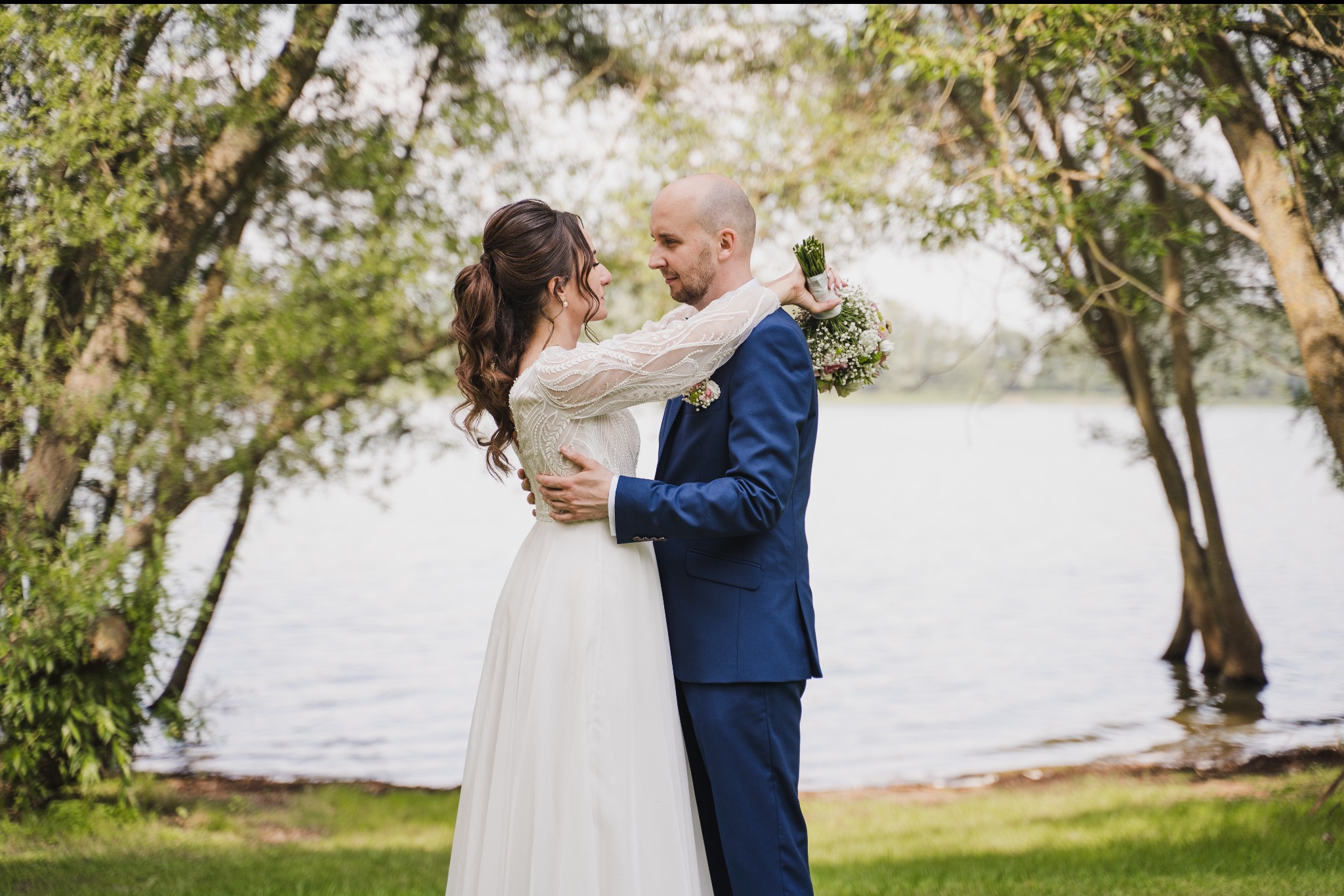 Para młoda w objęciach na tle jeziora i drzew, panna młoda w sukni z koronkowymi rękawami, pan młody w granatowym garniturze z butonierką, trzyma bukiet ślubny.