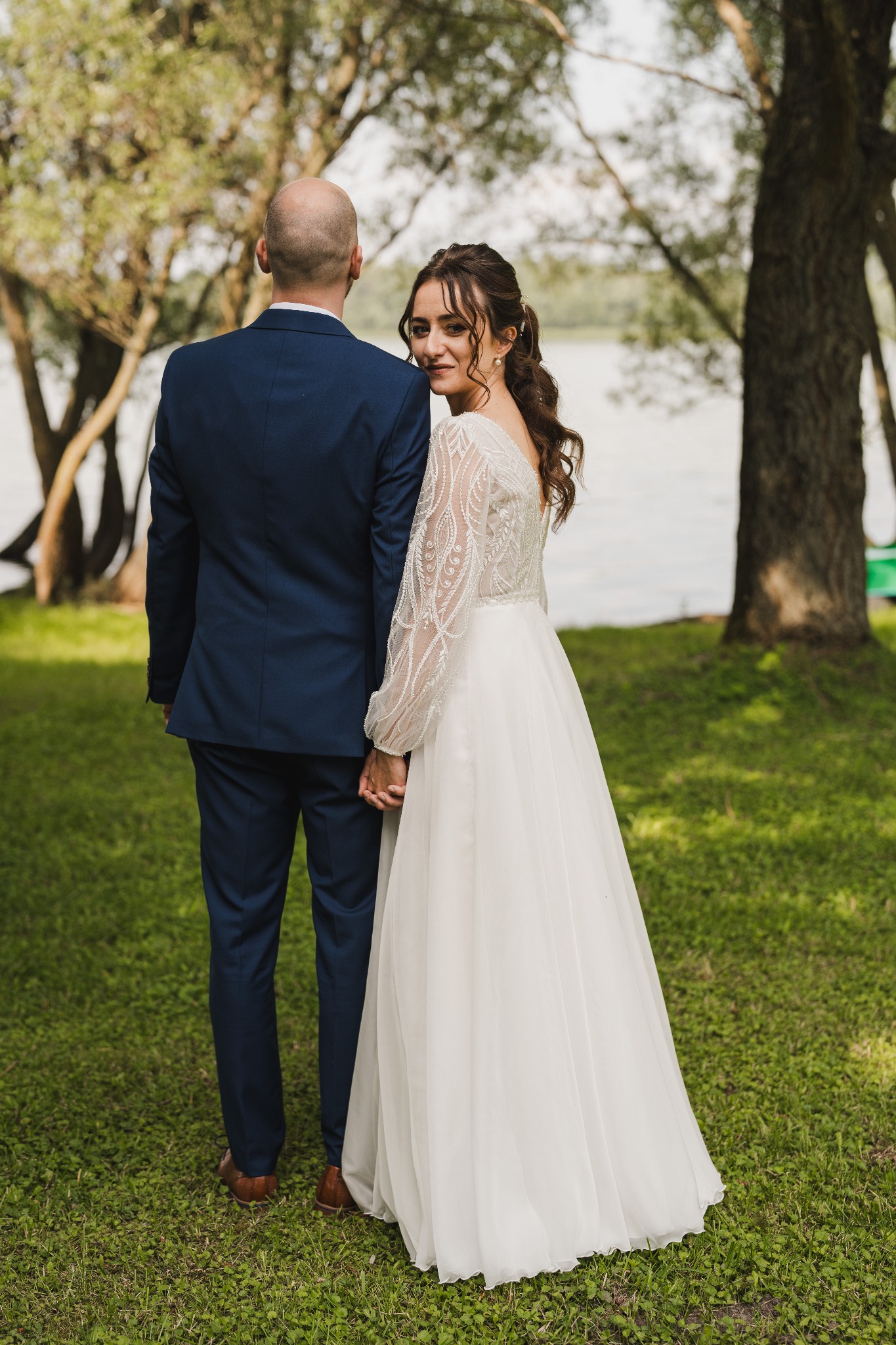 Para młoda trzymająca się za ręce, panna młoda odwraca się z uśmiechem, widok z tyłu na pana młodego w granatowym garniturze, zielona trawa i drzewa w tle.