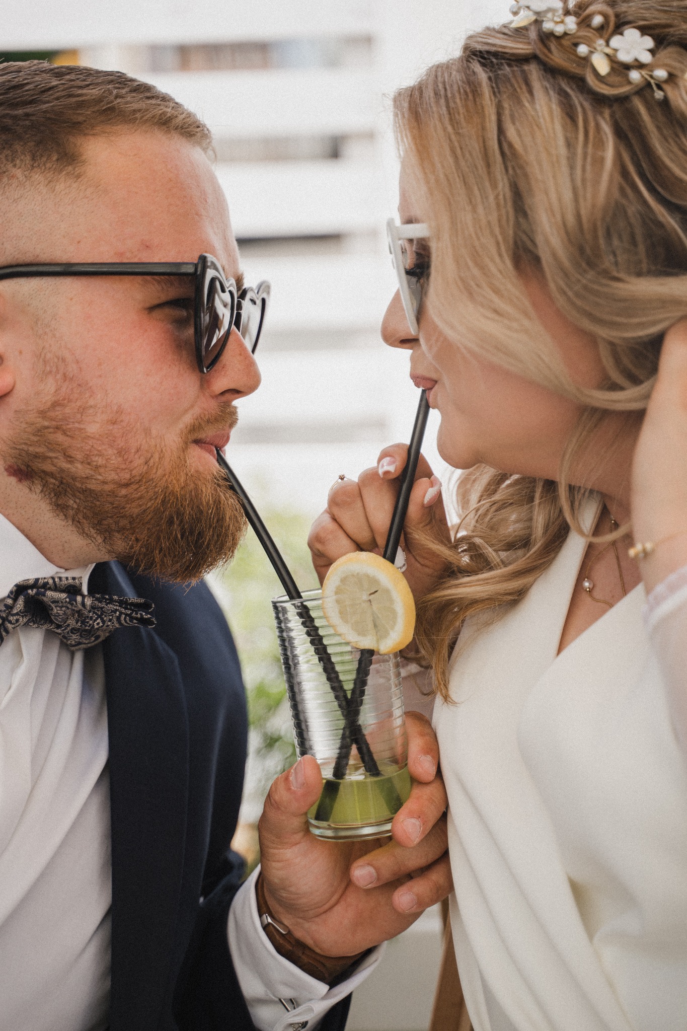 Para młoda w okularach pije drinka z dwóch słomek; panna młoda ma wpięte kwiaty we włosy, a pan młody ma muszkę.