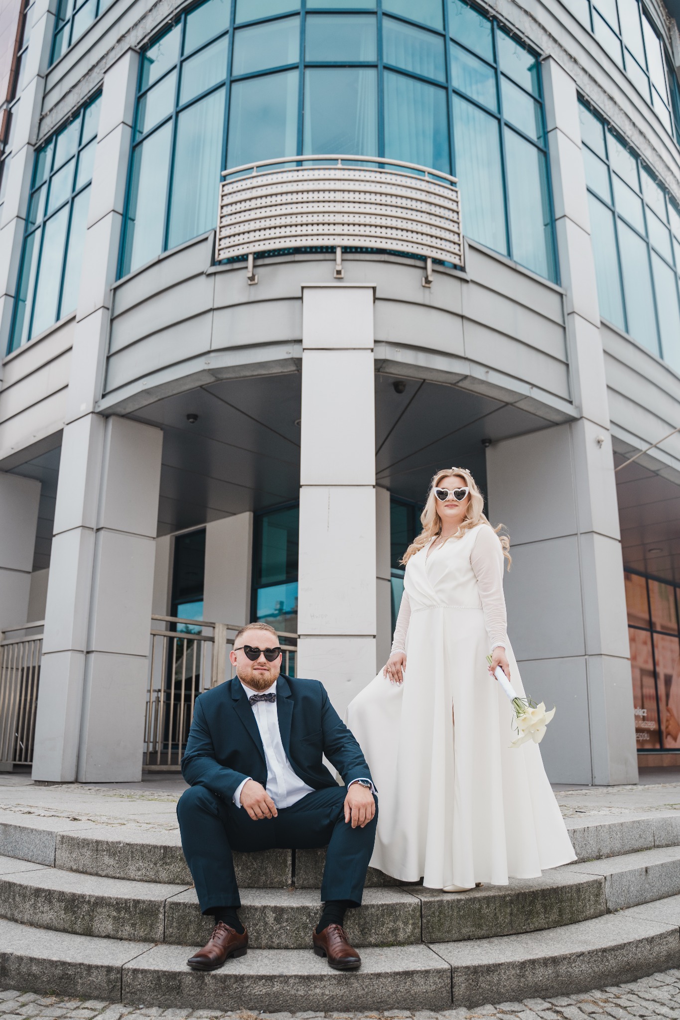 Para młoda w okularach w kształcie serc pozuje na schodach nowoczesnego budynku z przeszkloną fasadą.