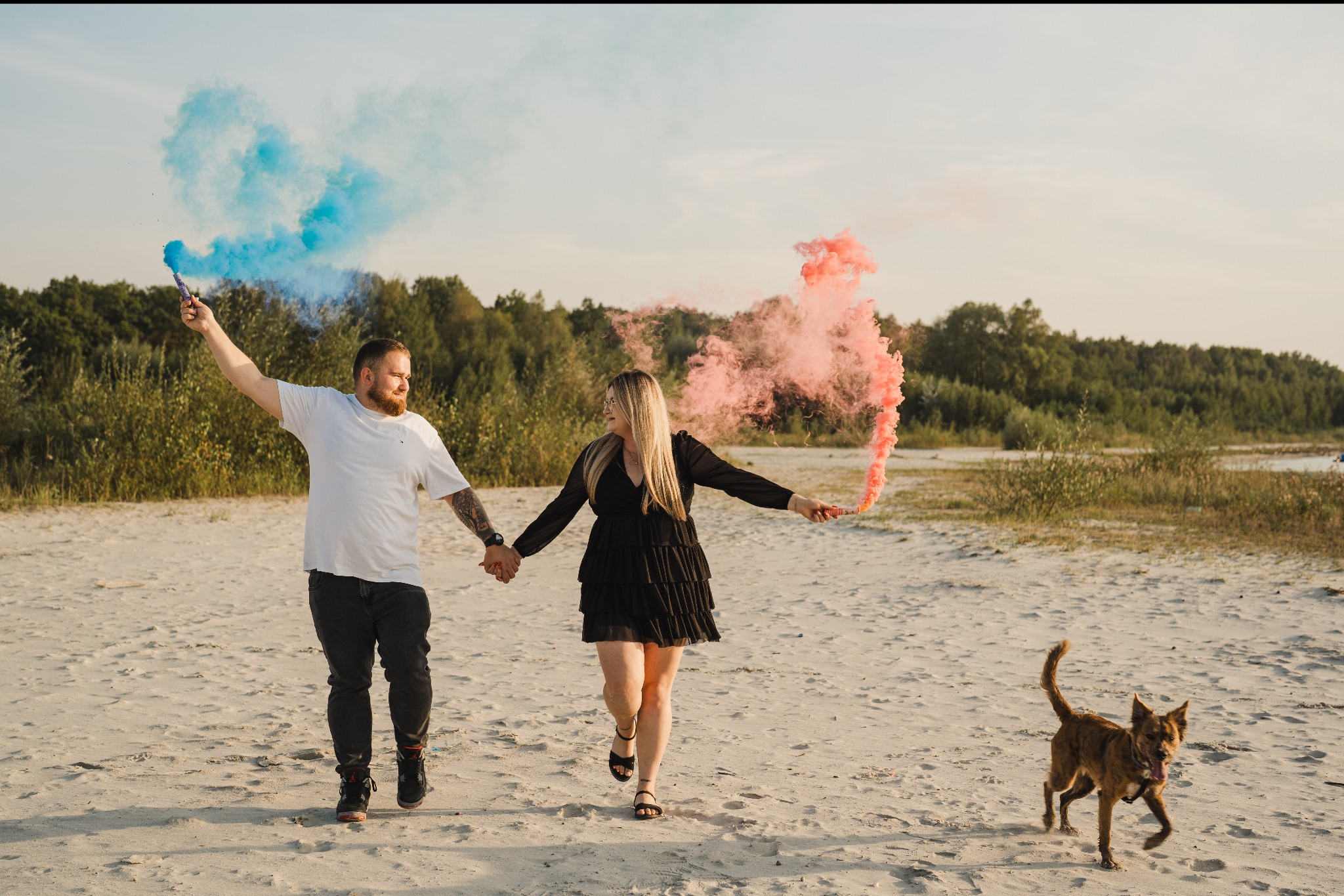Para trzymająca się za ręce, biegnąca po piaszczystej plaży z kolorowymi dymami, z psem biegnącym obok, w tle drzewa.