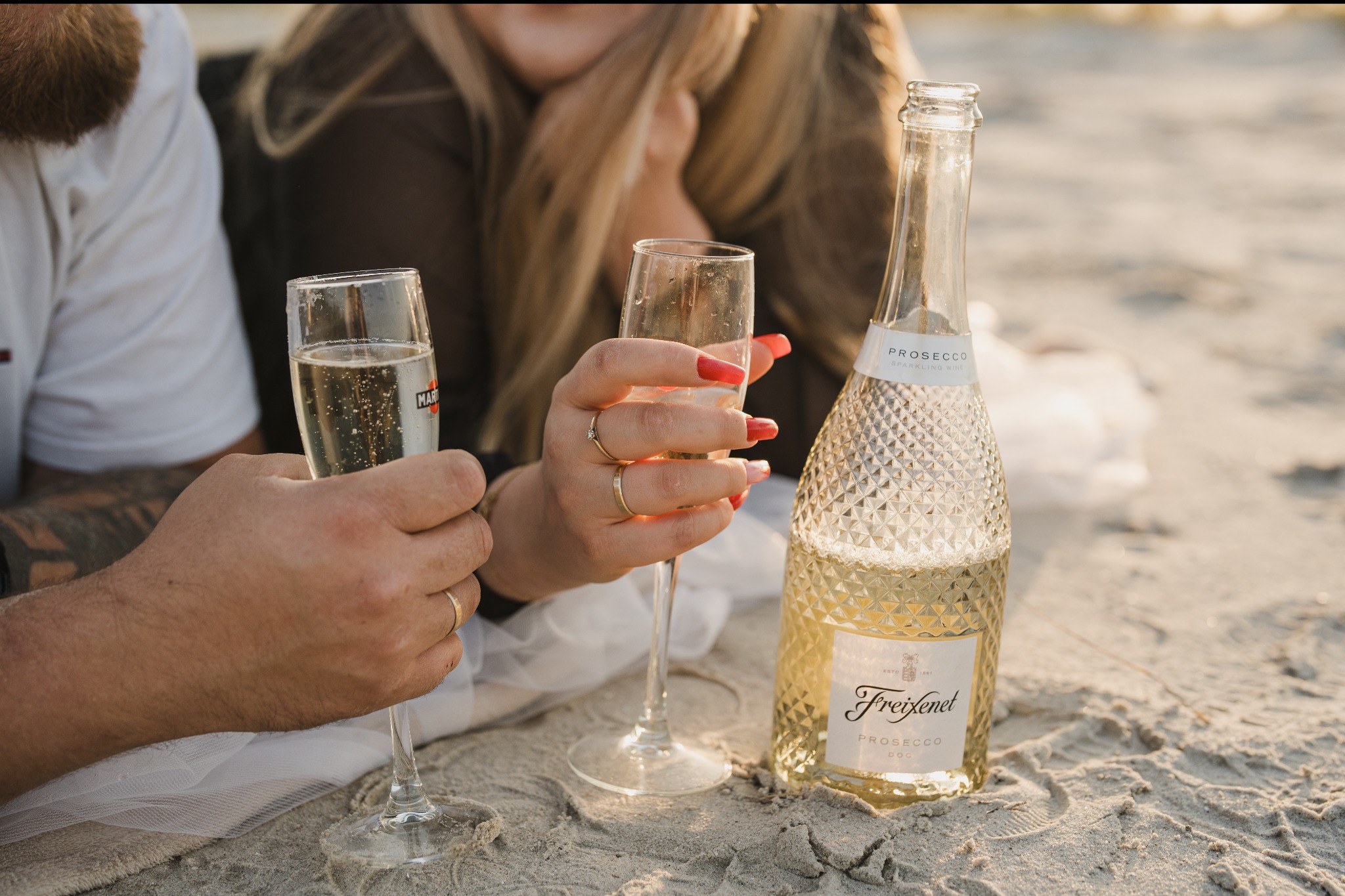 Para w plenerze na plaży, trzymająca kieliszki z Martini Prosecco i butelkę Freixenet Prosecco, z widocznymi pierścionkami na palcach i czerwonymi paznokciami u kobiety.