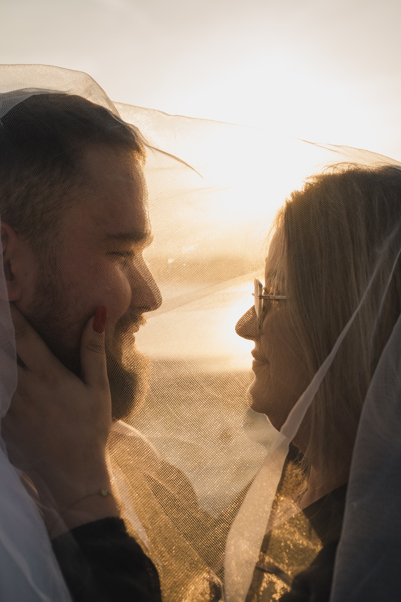 Profil młodej pary, częściowo przesłoniętej przez delikatny, prześwitujący materiał w ciepłym świetle zachodzącego słońca; kobieta w okularach patrzy na mężczyznę z brodą, którego dłoń dotyka jej...