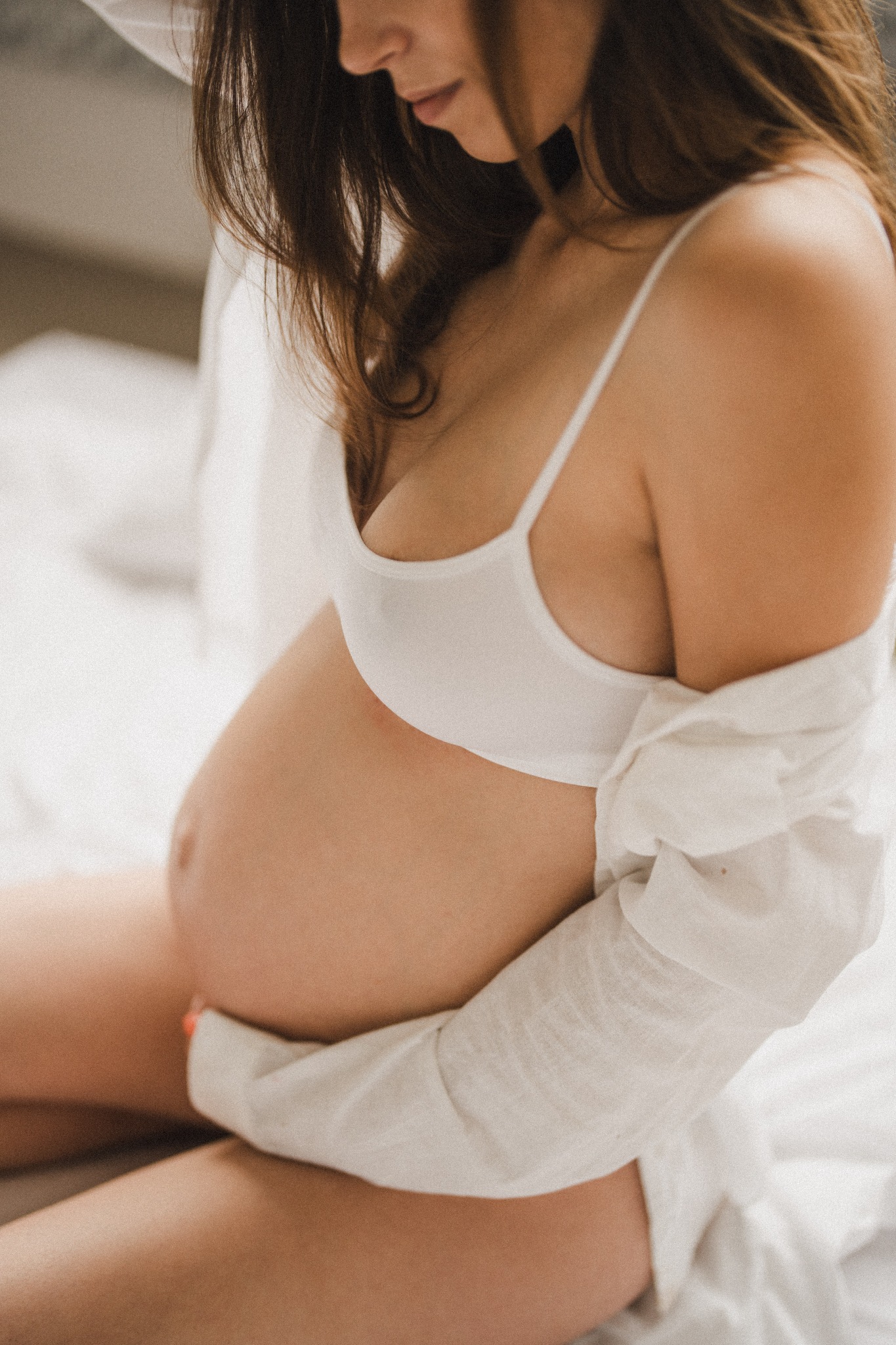 Kobieta w bieliźnie i rozpiętej koszuli obejmuje ciążowy brzuch, delikatne światło, neutralne tło.