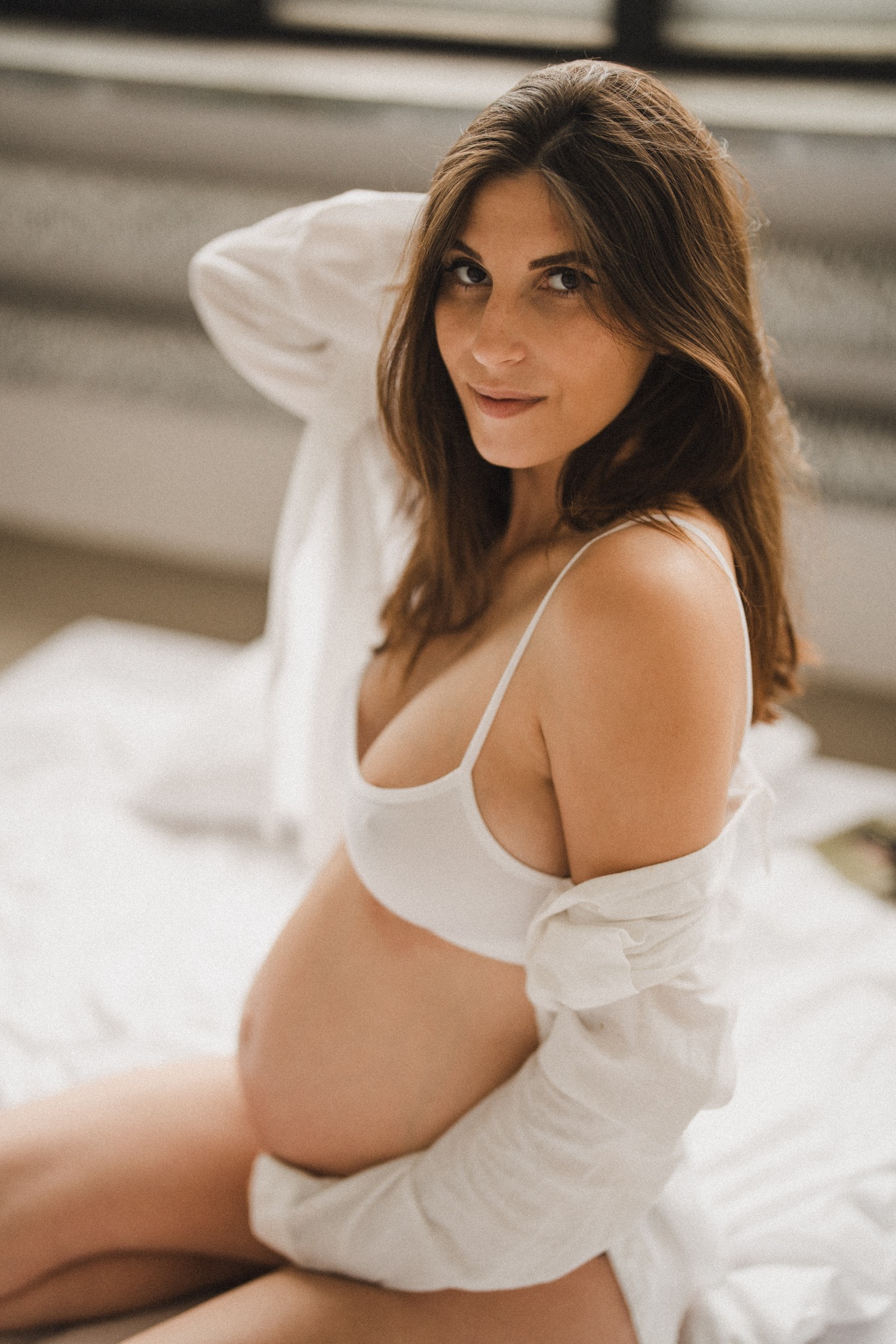 Kobieta w ciąży w białej bieliźnie i rozpiętej koszuli siedzi na białej pościeli, spoglądając w obiektyw z lekkim uśmiechem.