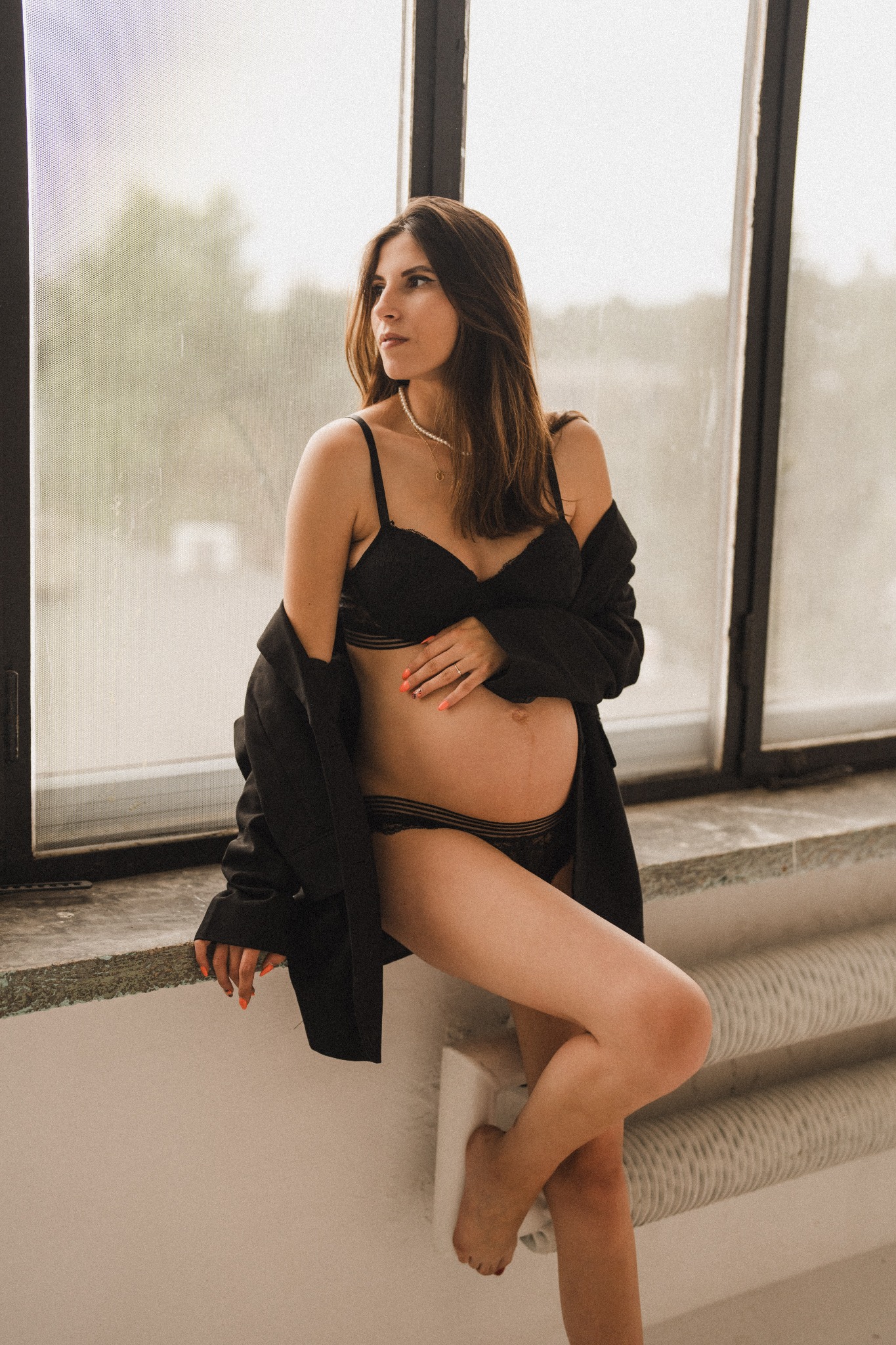 Kobieta w czarnej bieliźnie i marynarce, z ręką na ciążowym brzuchu, pozuje przy oknie w stonowanej kolorystyce.