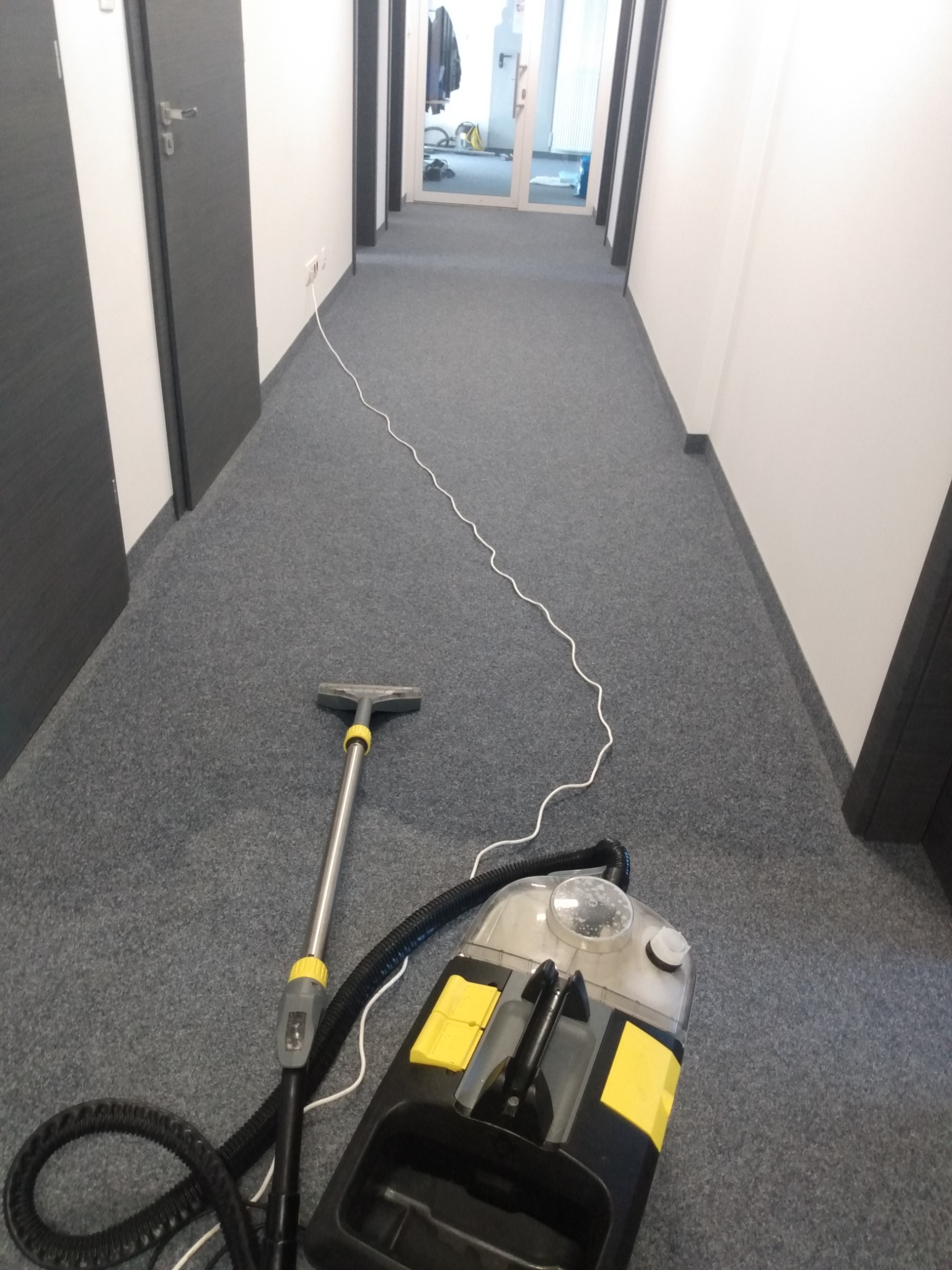 Długi korytarz biurowy z szarym dywanem, na którym stoi profesjonalny odkurzacz piorący z żółtymi akcentami i rozciągniętym kablem zasilającym.