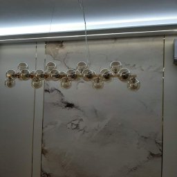 14 - Nowoczesny żyrandol z kulistymi kloszami w odcieniach bursztynu, zawieszony na tle ściany z marmurowym wzorem i listwą LED pod sufitem.