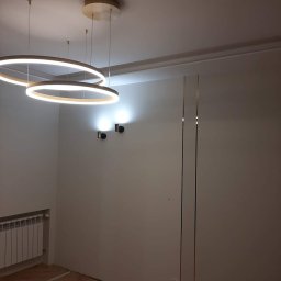 14 - Pokój z trzema okrągłymi lampami LED zwisającymi z sufitu, dwoma kinkietami na ścianie, grzejnikiem żeberkowym i parkietem ułożonym w jodełkę.