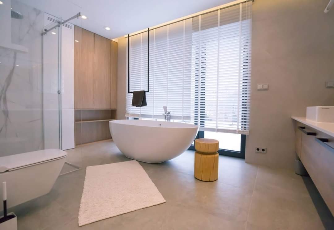 Nowoczesna łazienka z wolnostojącą wanną, drewnianym stołkiem i marmurową kabiną prysznicową, oświetlona światłem dziennym wpadającym przez żaluzje.