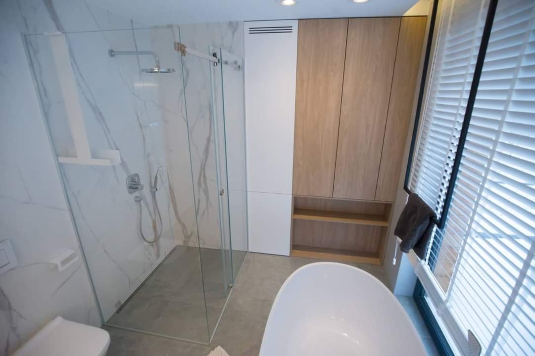 Nowoczesna łazienka z kabiną prysznicową typu walk-in, marmurowymi płytkami na ścianie, zabudową meblową z drewnianymi frontami i wolnostojącą białą wanną.