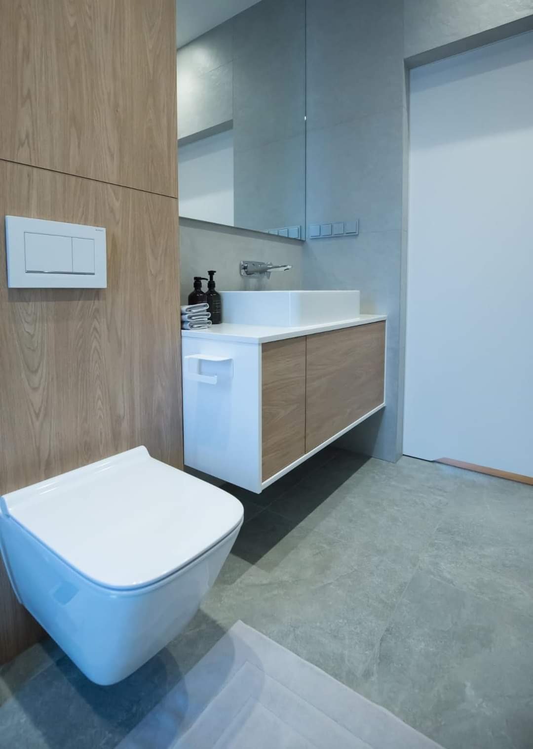 Nowoczesna łazienka z podwieszaną toaletą, szafką pod umywalką z drewnianymi frontami i szarą płytką podłogową.