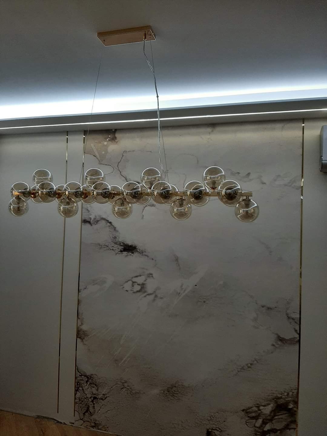 Nowoczesny żyrandol z kulistymi kloszami w odcieniach bursztynu, zawieszony na tle ściany z marmurowym wzorem i listwą LED pod sufitem.