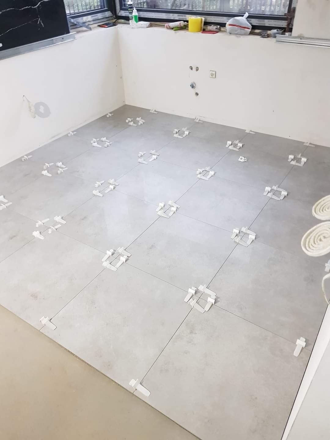 Szare płytki podłogowe w trakcie układania, z białymi krzyżykami dystansowymi, widoczne okno i fragment ściany z instalacją hydrauliczną.