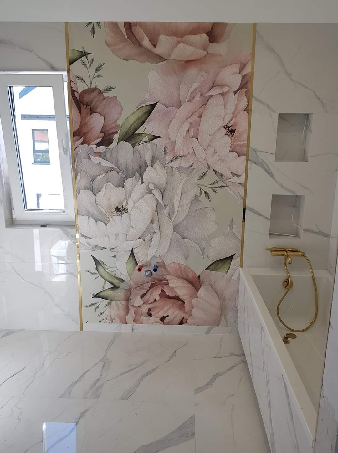 Wykończona łazienka z marmurowymi płytkami, dekoracyjną tapetą w kwiaty z złotymi listwami, białą wanną i złotą armaturą oraz wnękami w ścianie.