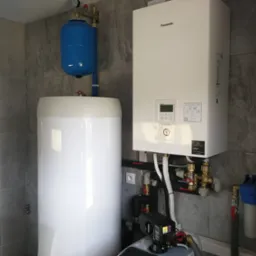 Biała pompa ciepła Panasonic Aquarea Smart Cloud zamontowana na szarej, kafelkowanej ścianie, z niebieskim zbiornikiem wyrównawczym i białym zasobnikiem ciepłej wody użytkowej obok, widoczne rury...