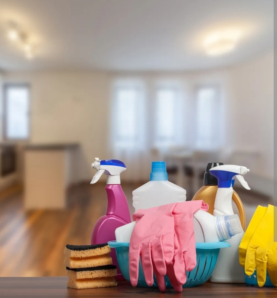 Zestaw środków czystości: gąbki, rękawice różowe i żółte, spryskiwacze i butelki w niebieskim koszyku na tle rozmytego wnętrza mieszkania.