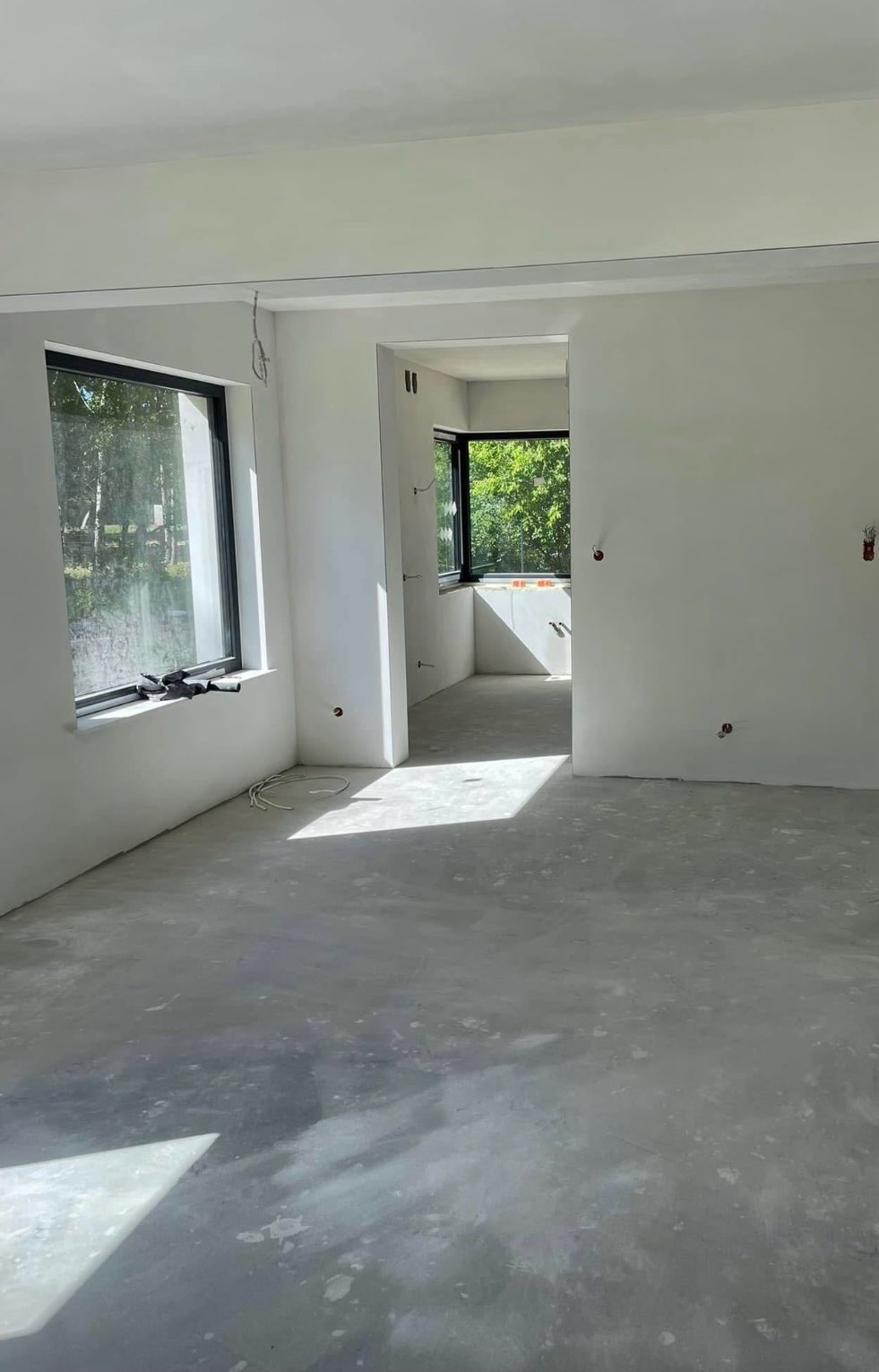 Wnętrze po wylewce anhydrytowej: jasne pomieszczenie z dużymi oknami, świeżo wylana, szara posadzka, przygotowane ściany pod malowanie. Widoczne kable i puszki.