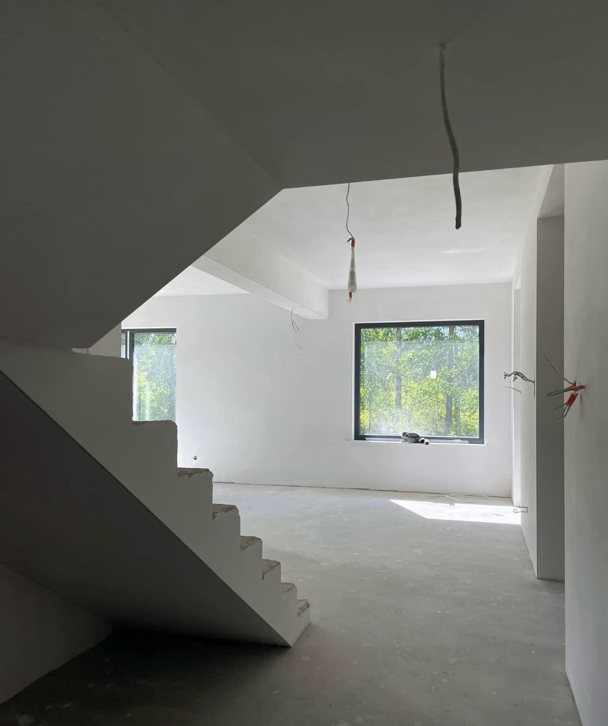 Wnętrze po tynkowaniu w Kamienicy Polskiej: białe ściany, schody i okno z widokiem na zieleń. Widoczne przewody elektryczne i surowa posadzka.