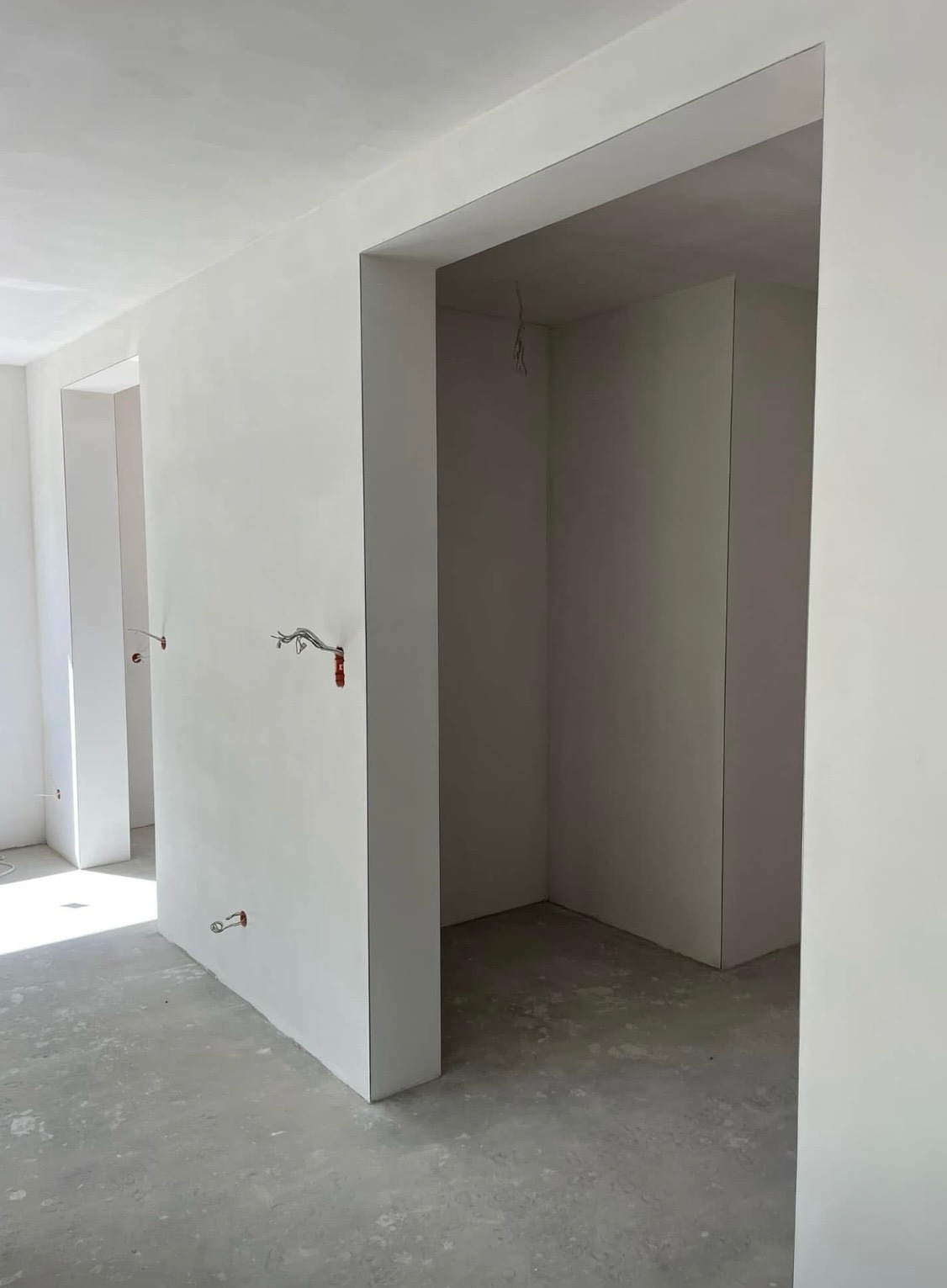 Wnętrze po gładziowaniu: białe ściany, sufit i wnęki drzwiowe, widoczne wystające przewody instalacji elektrycznej, surowa posadzka. Minimalistyczny efekt wykończenia.