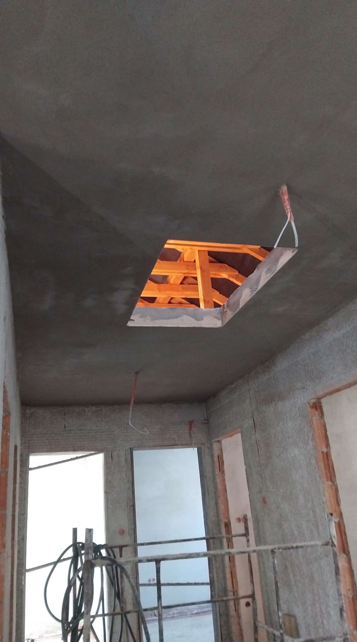 Szara, surowa powierzchnia sufitu z kwadratowym otworem ukazującym drewnianą konstrukcję dachu. W tle widoczne surowe ściany i otwory okienne/drzwiowe.