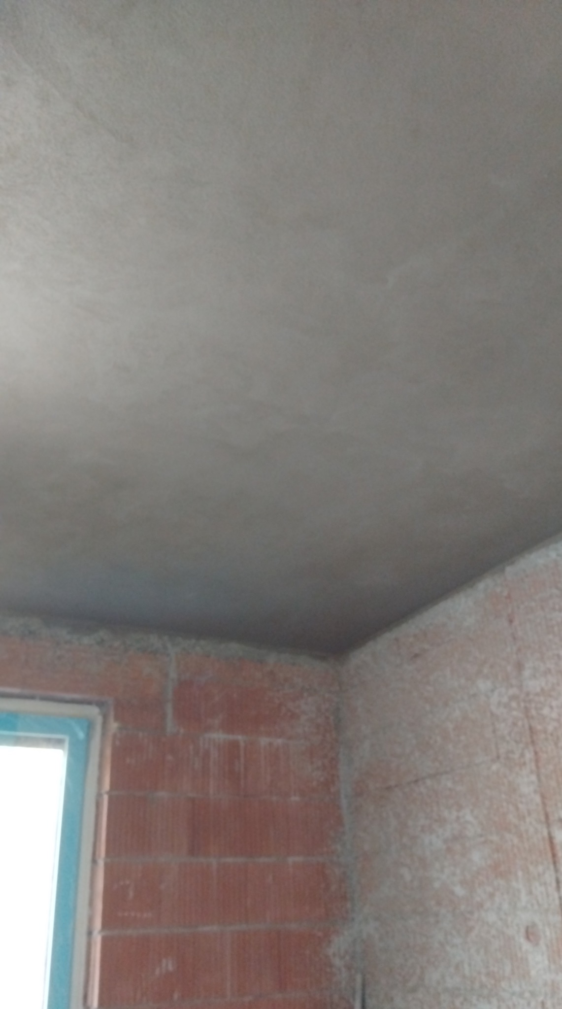 Świeżo położona, szara gładź na suficie i ścianach z czerwonej cegły. Widoczne okno z niebieską ramą. Nierównomierna faktura gładzi. Wnętrze w trakcie wykańczania.