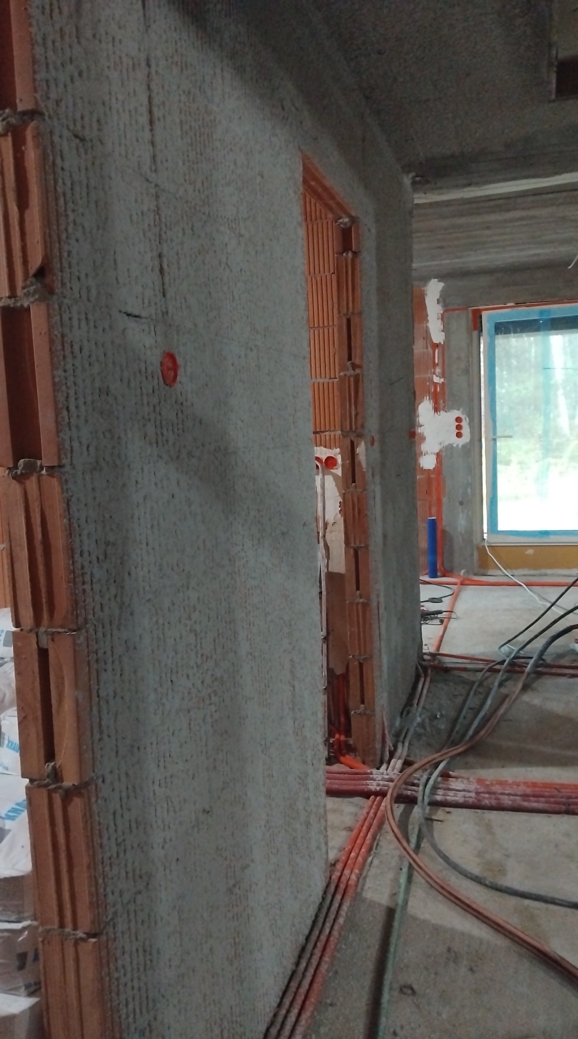 Ściana z tynkiem strukturalnym w trakcie prac wykończeniowych, widoczne instalacje elektryczne i fragment okna z niebieską ramą. Surowy stan wnętrza.