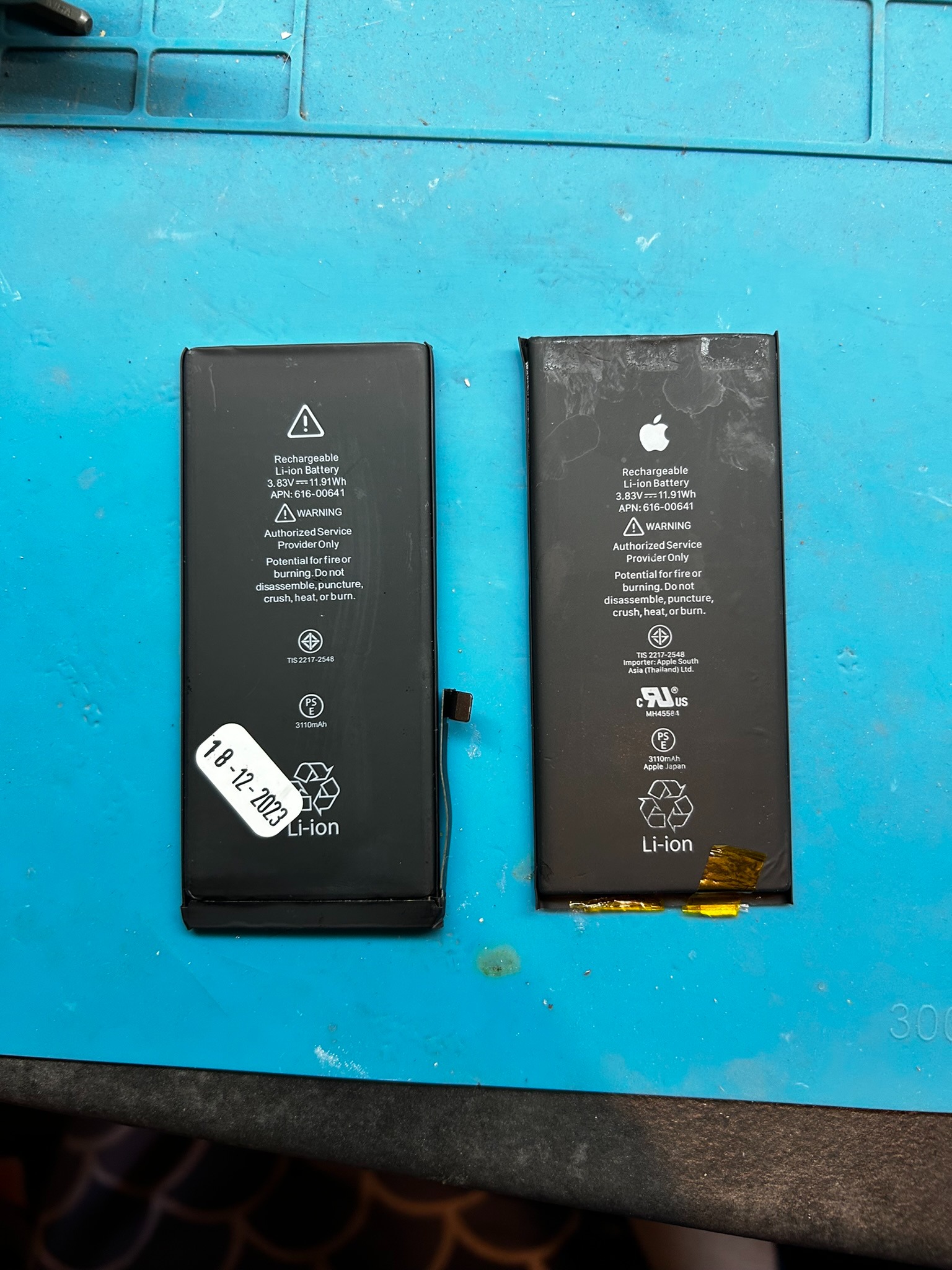 Dwie baterie litowo-jonowe do iPhone'a na niebieskiej macie serwisowej, jedna z naklejoną datą 18-12-2023, druga z uszkodzonym złączem.