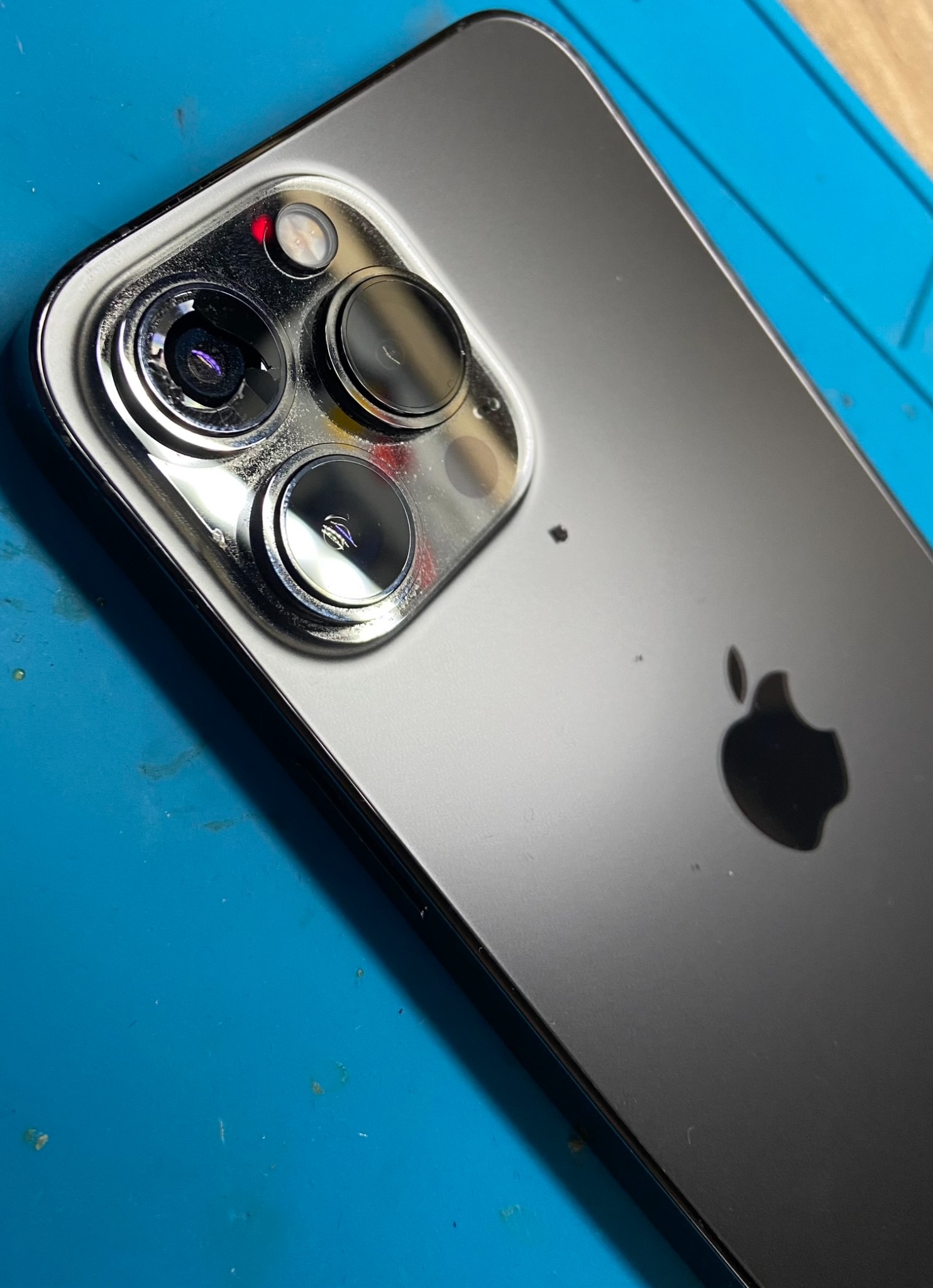 Widok z bliska na tylną obudowę szarego iPhone'a z trzema obiektywami aparatu i logo Apple, leżącego na niebieskiej macie serwisowej z widocznymi drobnymi zabrudzeniami.