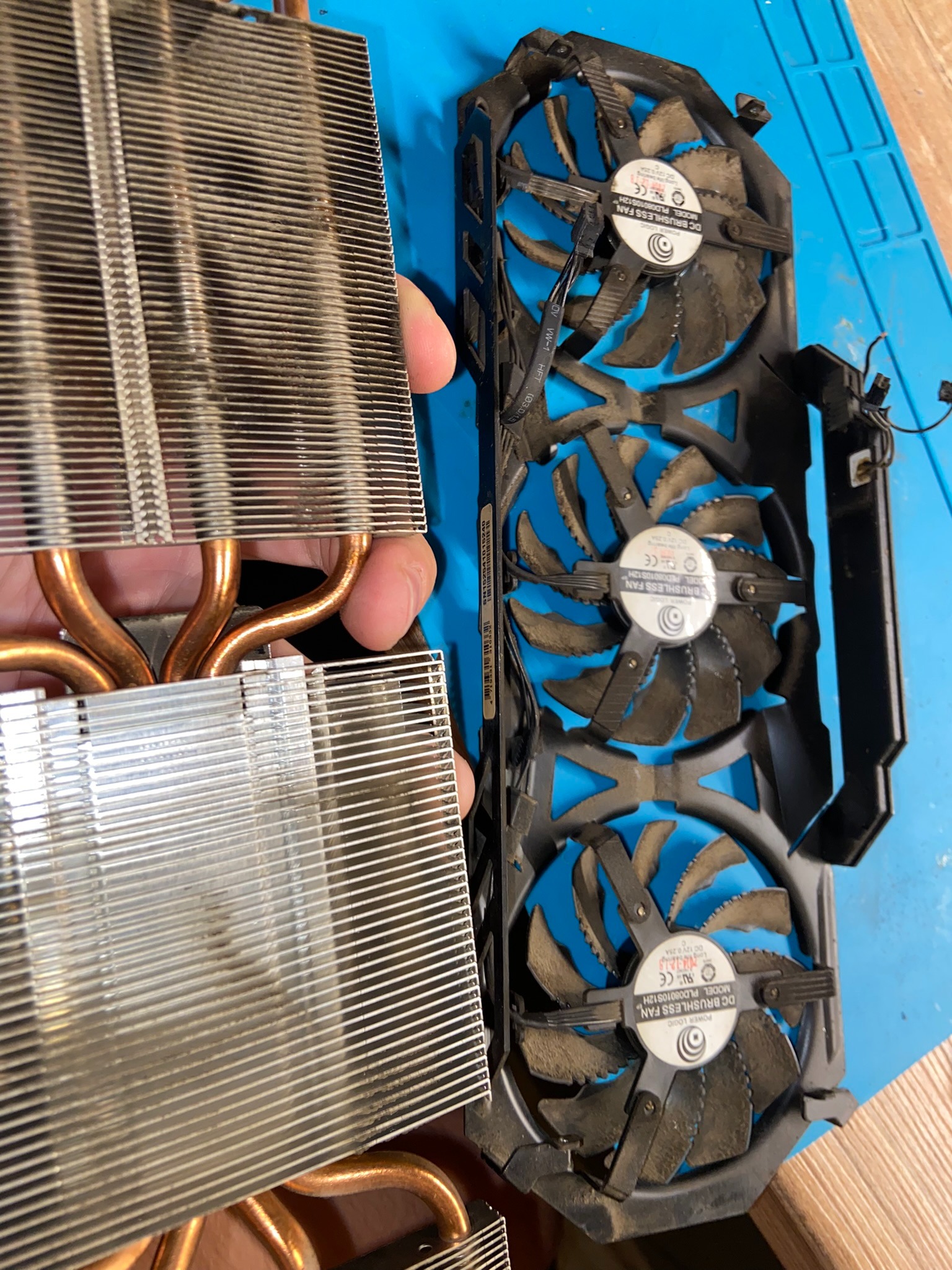 Brudna karta graficzna z trzema wentylatorami i odłączonym radiatorem z miedzianymi rurkami cieplnymi, leżąca na niebieskiej macie serwisowej.