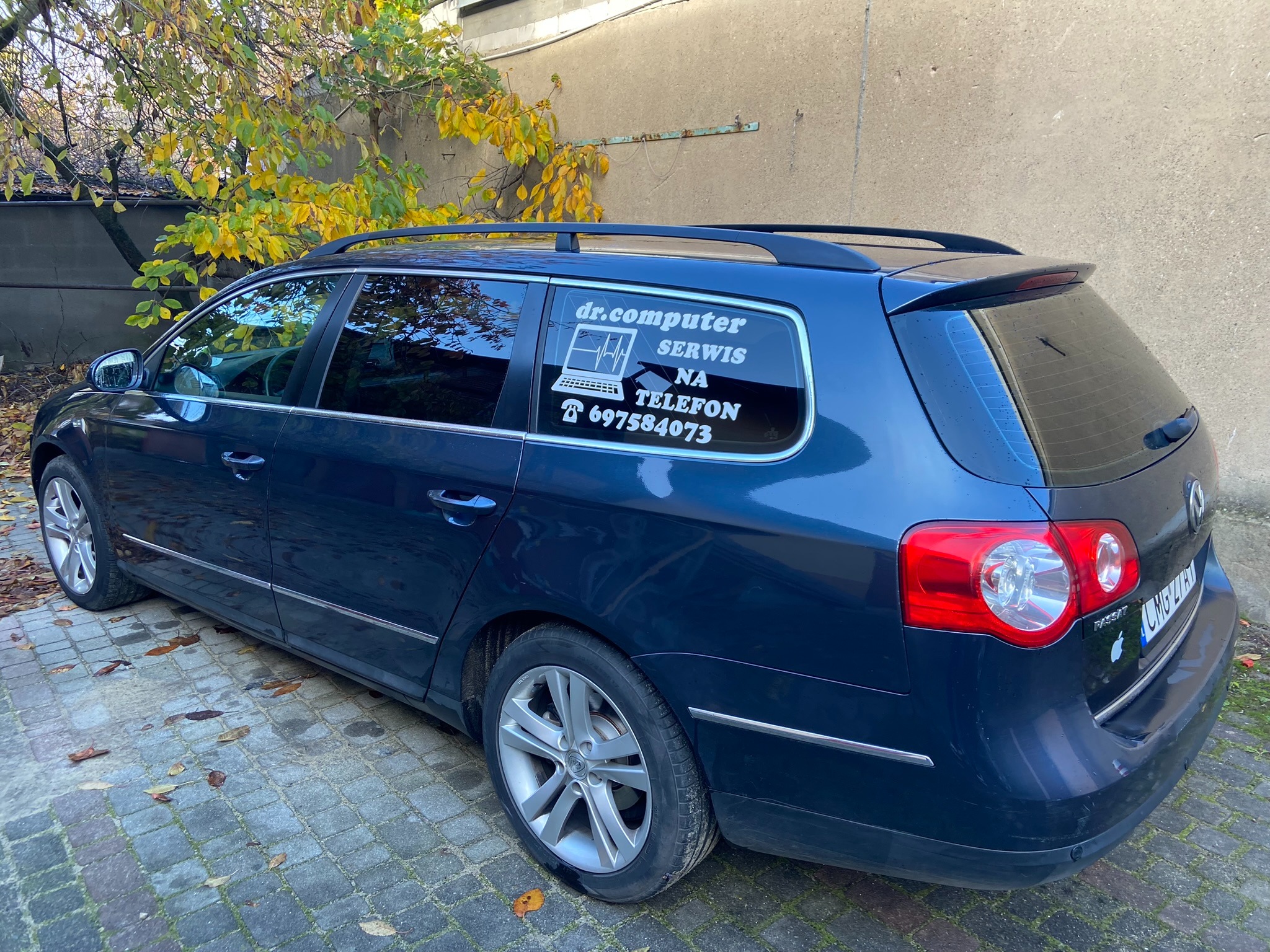 Niebieski samochód kombi z naklejką na szybie: 'dr.computer SERWIS NA TELEFON 697584073' i grafiką laptopa z wykresem EKG, zaparkowany na brukowanej powierzchni z jesiennymi liśćmi w tle.