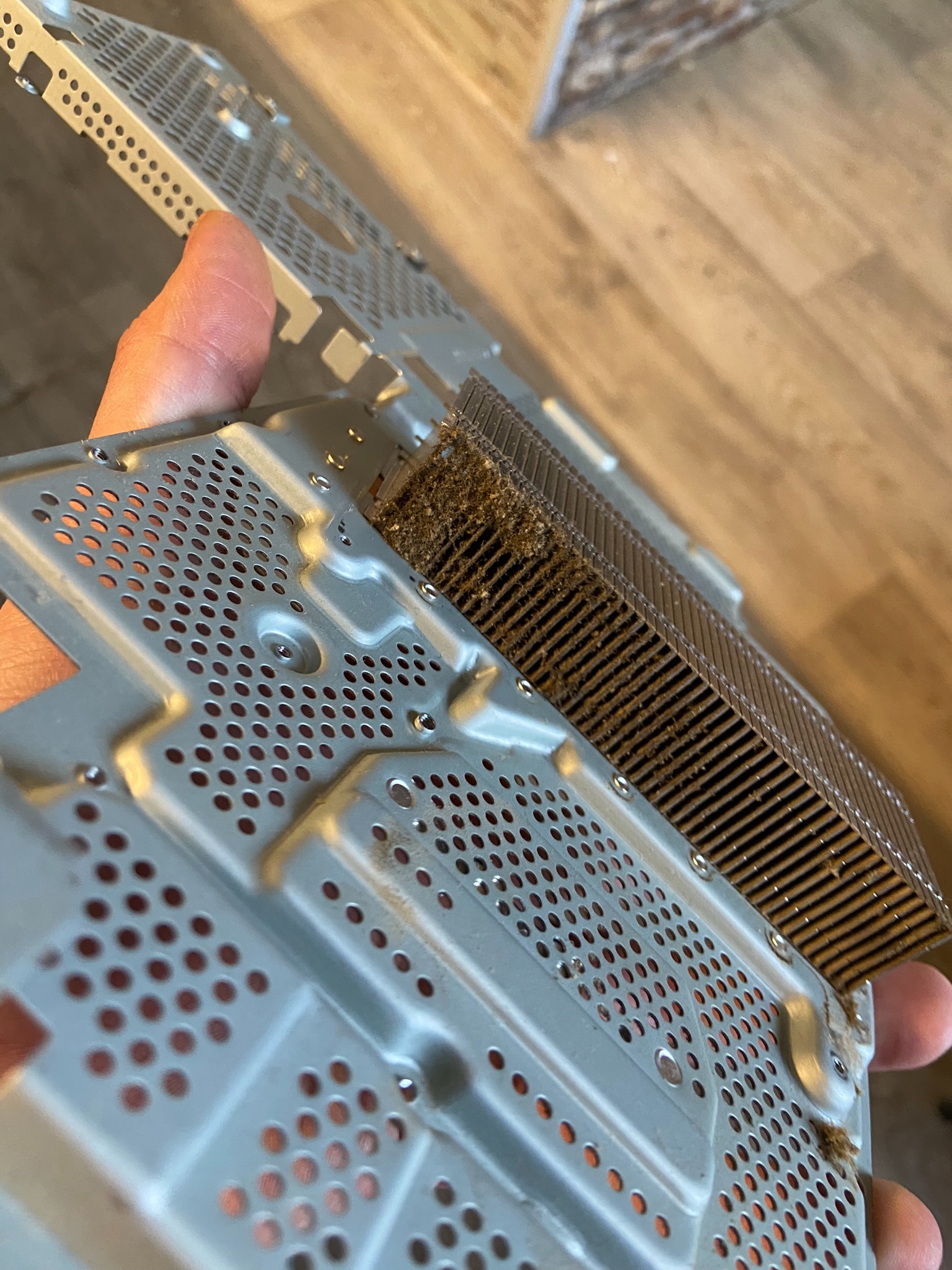 Brudny radiator procesora z kurzem i zanieczyszczeniami, wyjęty z obudowy komputera, trzymany w dłoni na tle drewnianej podłogi.