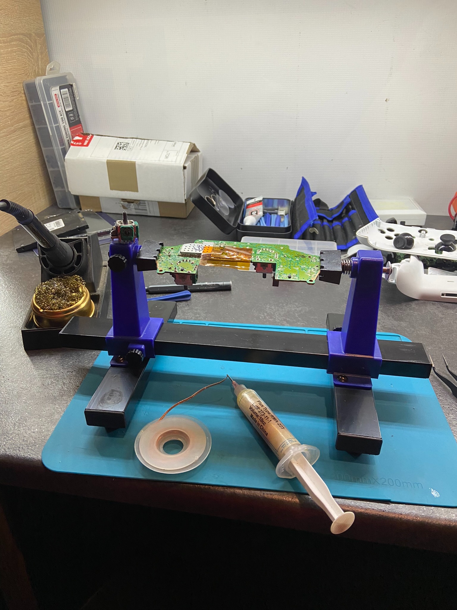 Płytka drukowana konsoli do gier zamocowana w uchwycie warsztatowym, obok lutownica, cyna, topnik w strzykawce oraz kontroler konsoli w tle.
